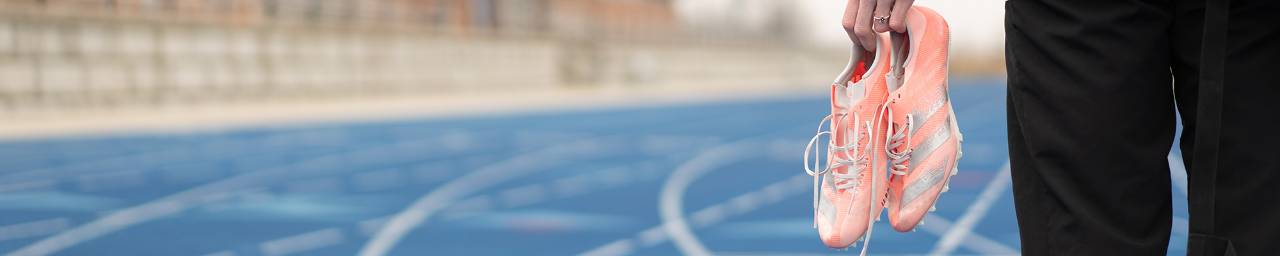 A close up of pink colored track cleats in the hands of an athlete while standing on a blue track.